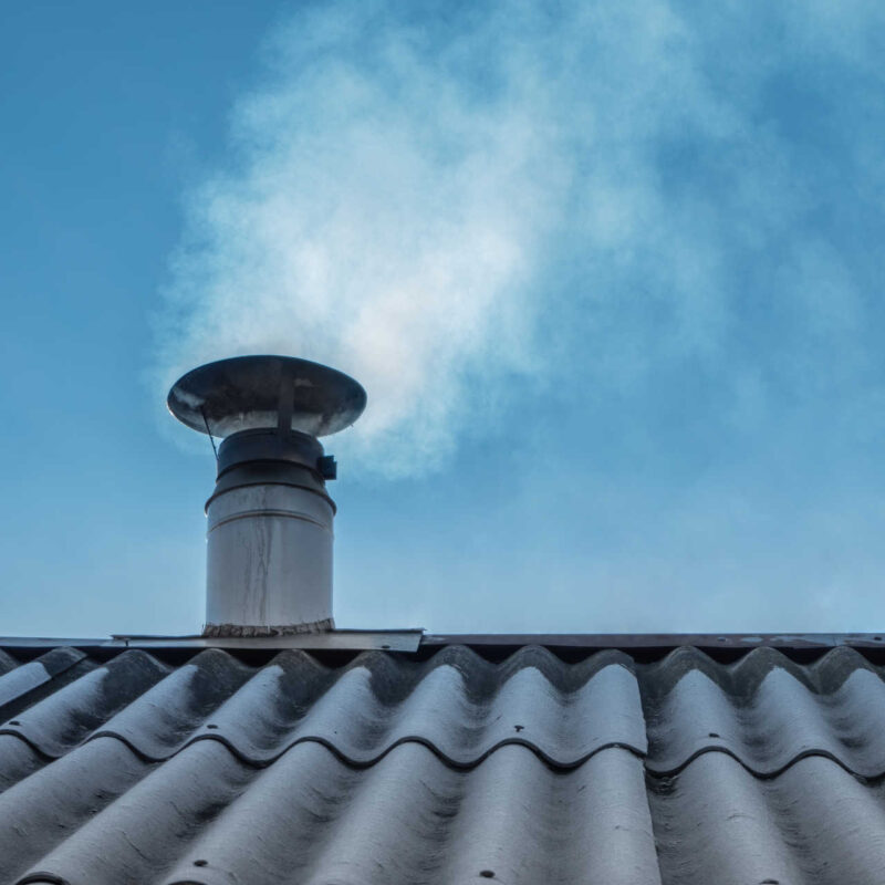 Aus einem Edelstahl-Kamin auf einem Dach steigt weißer Rauch in den blauen Himmel auf